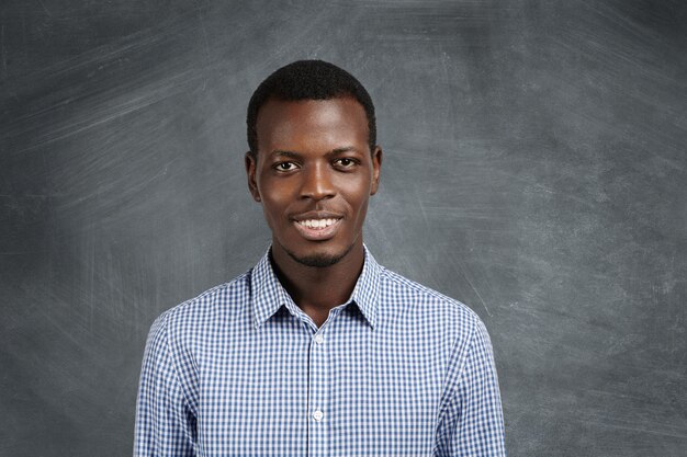 Retrato de um estudante atraente de pele escura, vestindo uma camisa xadrez com uma expressão confiante e alegre, em pé na parede do quadro