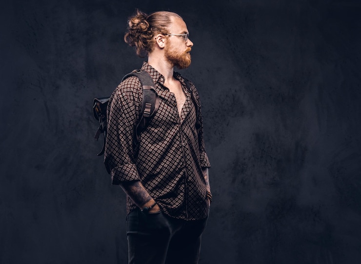 Foto grátis retrato de um estudante hipster ruiva de óculos vestido com uma camisa marrom, segura uma mochila, posando em um estúdio. isolado em um fundo escuro.
