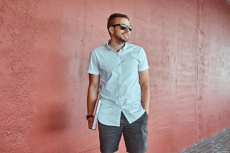 Foto grátis retrato de um freelancer elegante bonito feliz em óculos de sol vestido com uma camisa branca e shorts detém um computador portátil em pé debaixo de uma ponte contra uma parede, olhando para longe.
