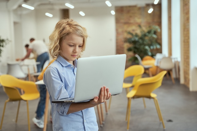 Retrato de um garotinho ocupado segurando e usando um laptop enquanto posava em uma sala de aula