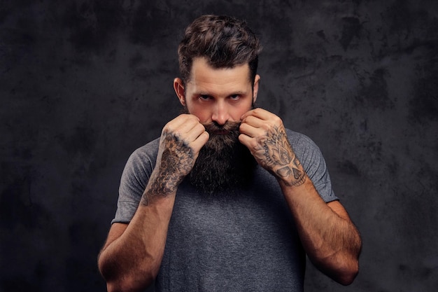 Retrato de um hipster tatuado com barba cheia e corte de cabelo elegante, vestido com uma camiseta cinza, olhando para a câmera, fica em um estúdio em um fundo escuro.