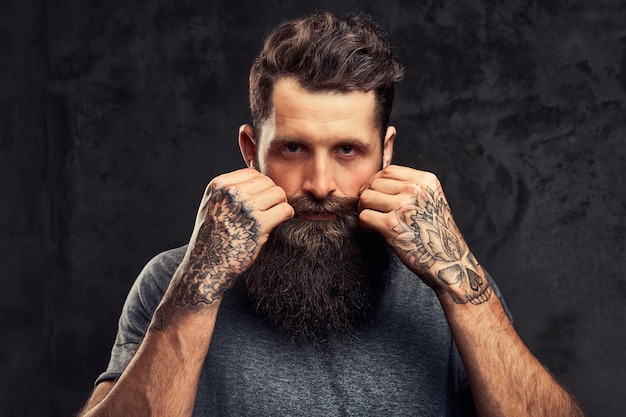 Retrato de um hipster tatuado com barba cheia e corte de cabelo elegante, vestido com uma camiseta cinza, olhando para a câmera, fica em um estúdio em um fundo escuro.