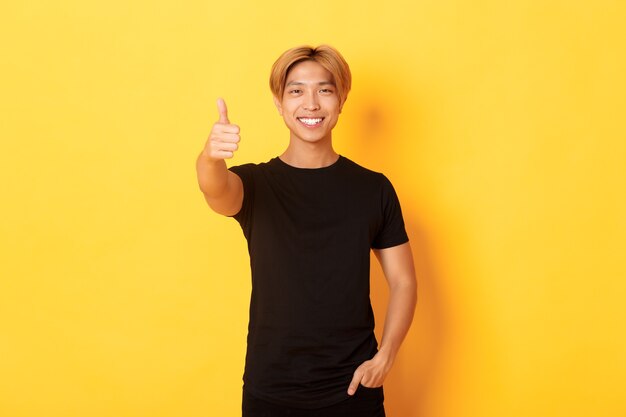 Retrato de um homem asiático satisfeito com cabelo loiro, em pé sobre a parede amarela e mostrando o polegar para cima