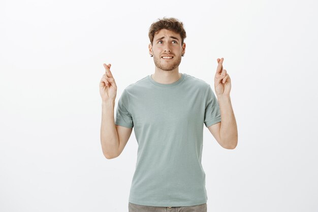 Retrato de um homem atraente e impaciente, nervoso, com brincos e camiseta pretos, levantando os dedos cruzados, mordendo o lábio e franzindo a testa enquanto olha para cima e espera que o desejo se torne realidade