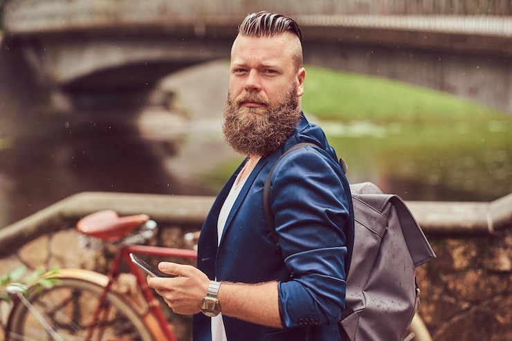 Foto grátis retrato de um homem barbudo com um corte de cabelo vestido com roupas casuais com uma mochila, de pé em um parque da cidade, usando um smartphone.