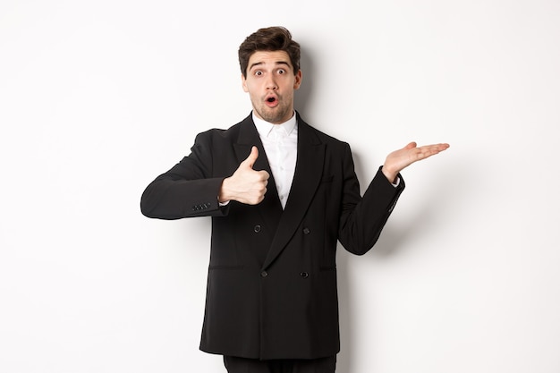 Retrato de um homem barbudo horrível em um terno formal, mostrando o polegar para cima e segurando o produto na mão sobre o espaço branco da cópia, recomendando o produto, de pé sobre um fundo branco