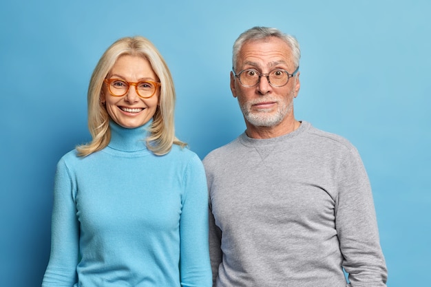 Retrato de um homem barbudo surpreso e uma mulher loira de meia-idade feliz, próximos um do outro, usando macacões casuais isolados sobre a parede azul do estúdio