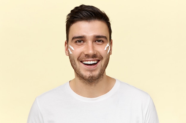 Retrato de um homem bonito de pele escura com a barba por fazer, tendo uma expressão facial alegre e enérgica, olhando para a câmera com um sorriso largo e radiante com listras de creme branco nas bochechas