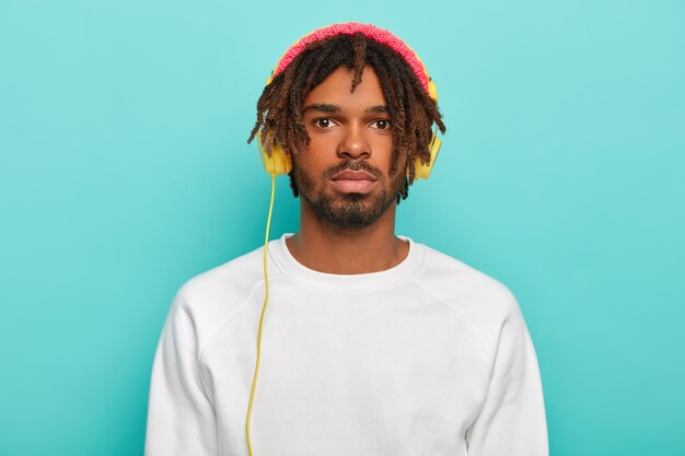 Retrato de um homem bonito e confiante com dreadlocks, usa fones de ouvido conectados a algum dispositivo, ouve música quente ou rádio, desfruta de um ambiente aconchegante, vestido com um suéter branco casual, capacete rosado