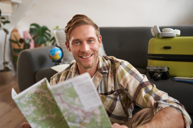 Retrato de um homem bonito e sorridente segurando um mapa sentado com uma mala na sala de estar indo para o exterior