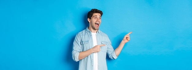 Foto grátis retrato de um homem bonito olhando e apontando os dedos deixados com emoção em pé contra o bac azul