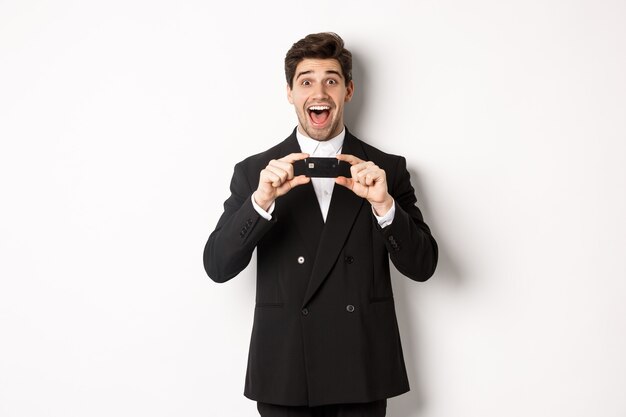 Retrato de um homem bonito surpreso em um terno preto, mostrando um cartão de crédito e recomendando um banco, em pé sobre um fundo branco
