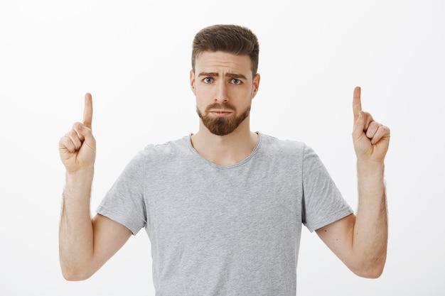 Retrato de um homem bonito, triste e bobo infeliz com barba e bigode franzindo a testa, fazendo um sorriso sombrio apontando para cima, expressando arrependimento ou ciúme em pé descontente contra a parede cinza