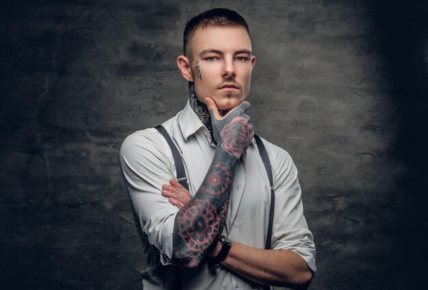 Retrato de um homem com uma tatuagem no rosto e braços, vestido com uma camisa branca sobre fundo cinza.