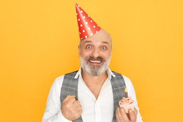 Retrato de um homem de negócios barbudo sênior de sucesso e atraente usando chapéu de cone, expressando empolgação, curtindo a festa de aniversário, segurando um bolinho com uma vela