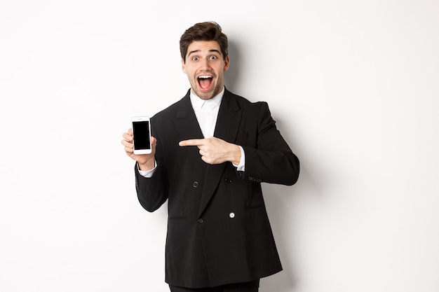 Retrato de um homem de negócios bonito de terno, apontando o dedo para a tela do celular, mostrando um anúncio, em pé sobre um fundo branco