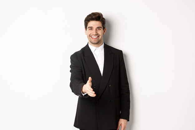 Retrato de um homem de negócios bonito em um terno preto, estendendo a mão para um aperto de mão, cumprimentando os parceiros de negócios e sorrindo, bem-vindo à empresa, em pé sobre um fundo branco