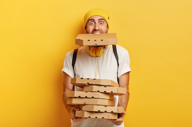 Retrato de um homem de pizza sobrecarregado contém muitos pacotes de papelão, um na boca, usa uma camiseta branca casual.