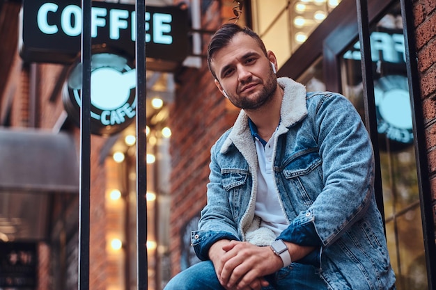 Retrato de um homem elegante, vestindo uma jaqueta jeans com fones de ouvido sem fio, segurando o café para viagem fora do café, olhando para a câmera.