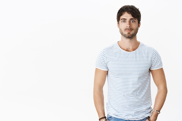 Retrato de um homem europeu atraente com olhos azuis e cabelos castanhos, cabelos ondulados sorrindo casualmente, segurando as mãos nos bolsos, encostado na parede cinza em uma camiseta listrada