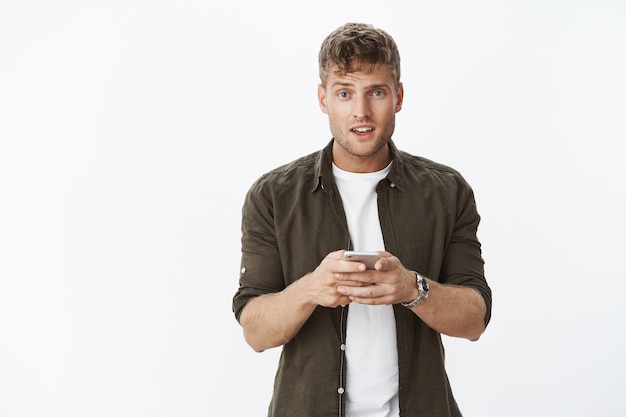 Foto grátis retrato de um homem europeu fofo confuso e distraído com cabelo loiro e cabeça erguida na frente, questionado quando interrompido por usar um smartphone sobre uma parede cinza