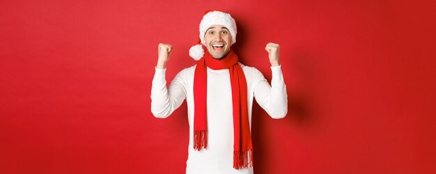 Retrato de um homem feliz e animado com chapéu de papai noel e lenço, regozijando-se e ganhando algo, comemorando o ano novo, em pé sobre fundo vermelho