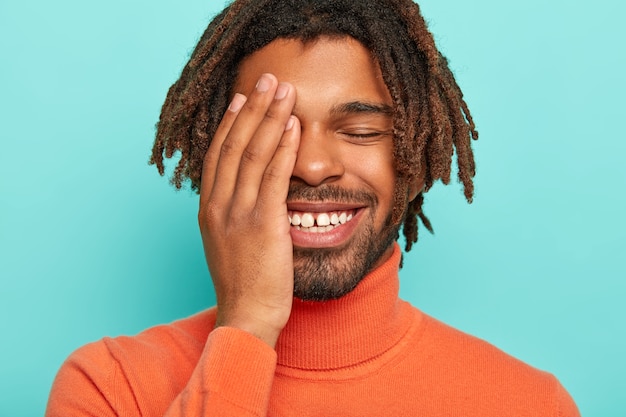 Retrato de um homem feliz e satisfeito com um sorriso largo, dentes brancos com pequenas lacunas, cobre o rosto com a palma da mão rindo de situações engraçadas