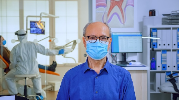 Retrato de um homem idoso no consultório odontológico, olhando na câmera, usando máscara facial, sentado na cadeira na clínica de estomatologia de sala de espera. Conceito de nova visita normal ao dentista em surto de coronavírus.