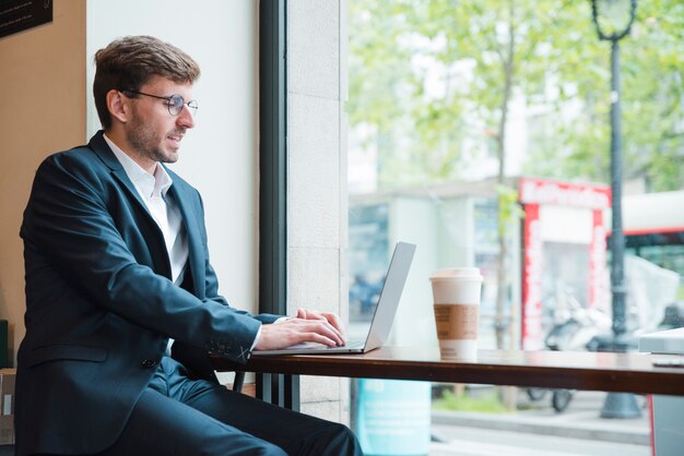Retrato, de, um, homem negócios, usando computador portátil, com, takeaway, xícara café, ligado, tabela, em, café