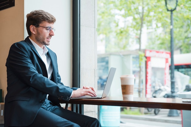 Retrato, de, um, homem negócios, usando computador portátil, com, takeaway, xícara café, ligado, tabela, em, café