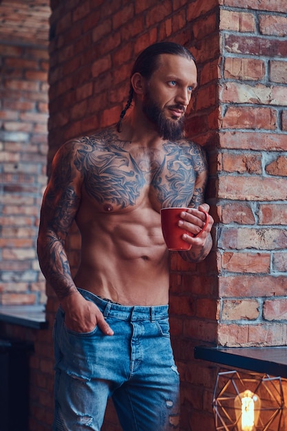 Retrato de um homem sem camisa tatuado com um corte de cabelo elegante e barba, bebe café da manhã, encostado em uma parede de tijolos em uma sala.