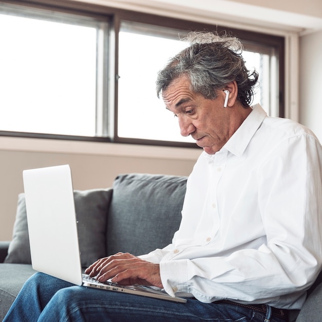 Retrato, de, um, homem sênior, sentar sofá, usando, bluetooth, fone ouvido, usando computador portátil