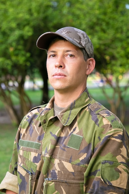 Retrato de um homem sério com uniforme de camuflagem militar em pé no parque,