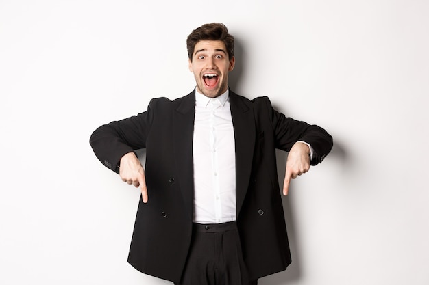 Retrato de um homem super feliz mostrando a oferta promocional de Natal, apontando o dedo para baixo e sorrindo espantado, vestindo terno formal de festa, em pé sobre um fundo branco