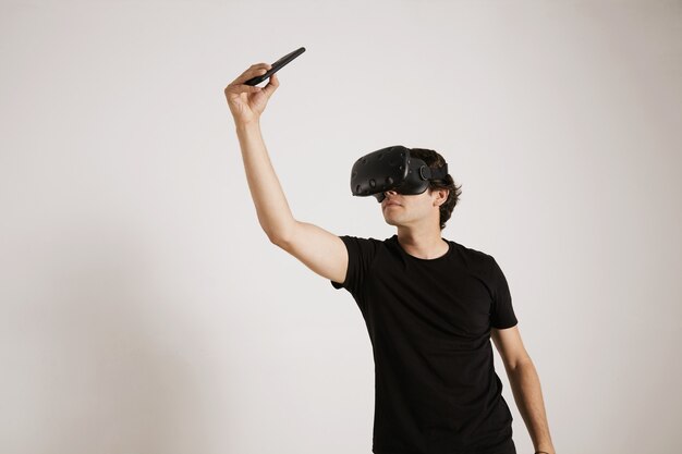 Retrato de um jogador com fone de ouvido de RV e camiseta preta em branco tirando uma selfie com seu smartphone