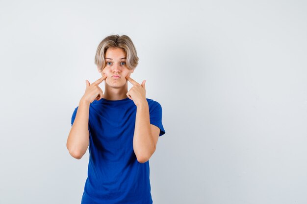 Retrato de um jovem adolescente apontando para as bochechas inchadas em uma camiseta azul e parecendo desapontado com a vista frontal