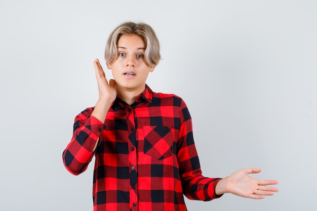 Retrato de um jovem adolescente com a mão perto da orelha, espalhando a palma da mão para o lado em uma camisa quadriculada e olhando a vista frontal perplexa