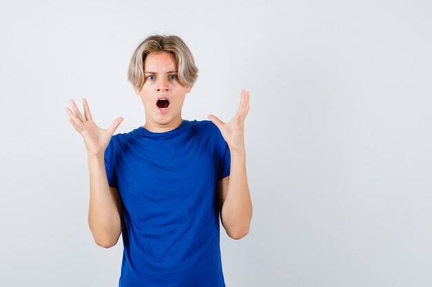 Retrato de um jovem adolescente levantando as mãos enquanto grita em uma camiseta azul e olhando a vista frontal apavorada