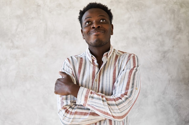 Retrato de um jovem afro-americano pensativo e sonhador em uma camisa listrada, abraçando-se e olhando para cima com uma expressão facial pensativa, sorrindo, sonhando acordado