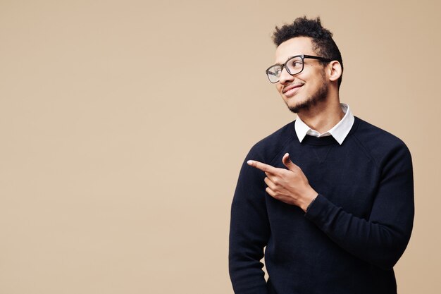 Retrato de um jovem bonito e sorridente homem afro de óculos, sorrindo e em pé com um gesto de mão aberta isolado na parede bege