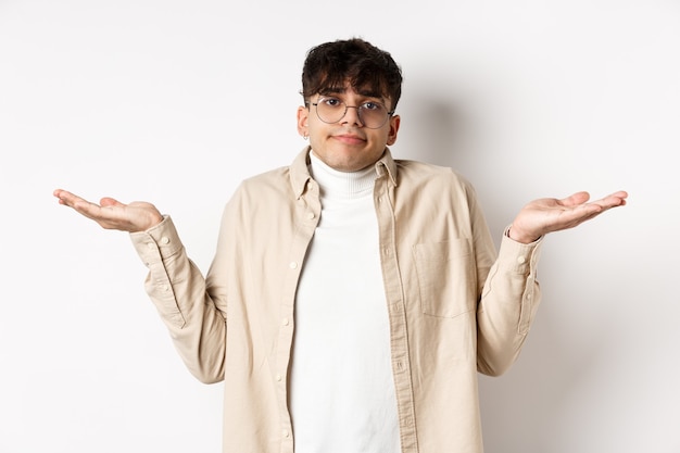 Retrato de um jovem bonito parecendo confuso, não sabe de nada, encolhendo os ombros e estendendo as mãos para os lados, sem noção, em pé sobre um fundo branco