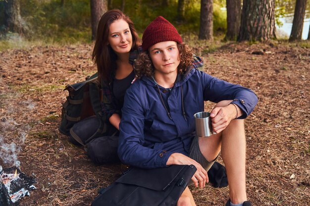 Retrato de um jovem casal - cara encaracolado bonito e garota encantadora sentados juntos na floresta. Conceito de viagens, turismo e caminhada.