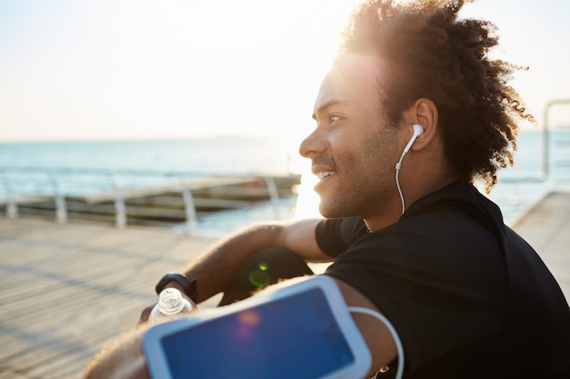 Retrato de um jovem desportista afro-americano sorridente com fones de ouvido em uma camiseta preta. descansando em um píer de madeira após uma corrida bem-sucedida. fazendo exercícios à beira-mar