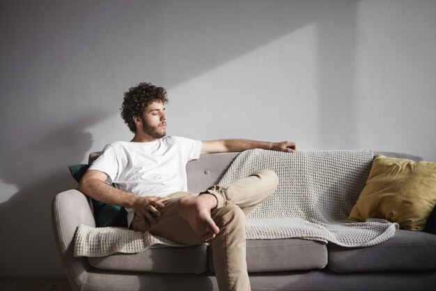 Retrato de um jovem elegante e atraente com a barba por fazer, vestindo uma camiseta branca e jeans bege, sentado descalço no sofá em casa, mantendo os olhos fechados, curtindo o sol, sentindo-se relaxado e despreocupado