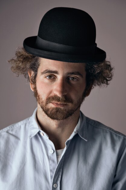 Retrato de um jovem judeu barbudo triste com cabelo encaracolado, vestindo um chapéu-coco preto engraçado e leve denim abotoar a t-shirt isolada em cinza claro.