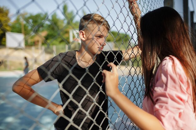 Retrato de um jovem legal parado na quadra de basquete e olhando pensativamente para uma menina bonita com cabelo escuro através da cerca de malha