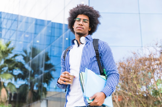 Retrato, de, um, jovem, macho, estudante americano africano, carregando saco, ligado, ombro, e, livros, em, mão, ficar, contra, universidade, predios