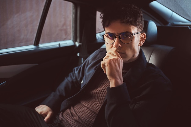 Retrato de um jovem pensativo de óculos sentado no banco de trás do carro.