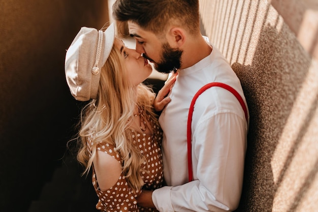 Retrato de um lindo casal romântico beijando ao ar livre no dia ensolarado garota de boné bege e vestido marrom olha nos olhos de seu amado namorado