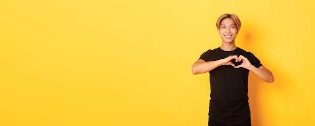 Retrato de um lindo e bonito homem asiático loiro mostrando gesto cardíaco e sorrindo em pé amarelo backgr